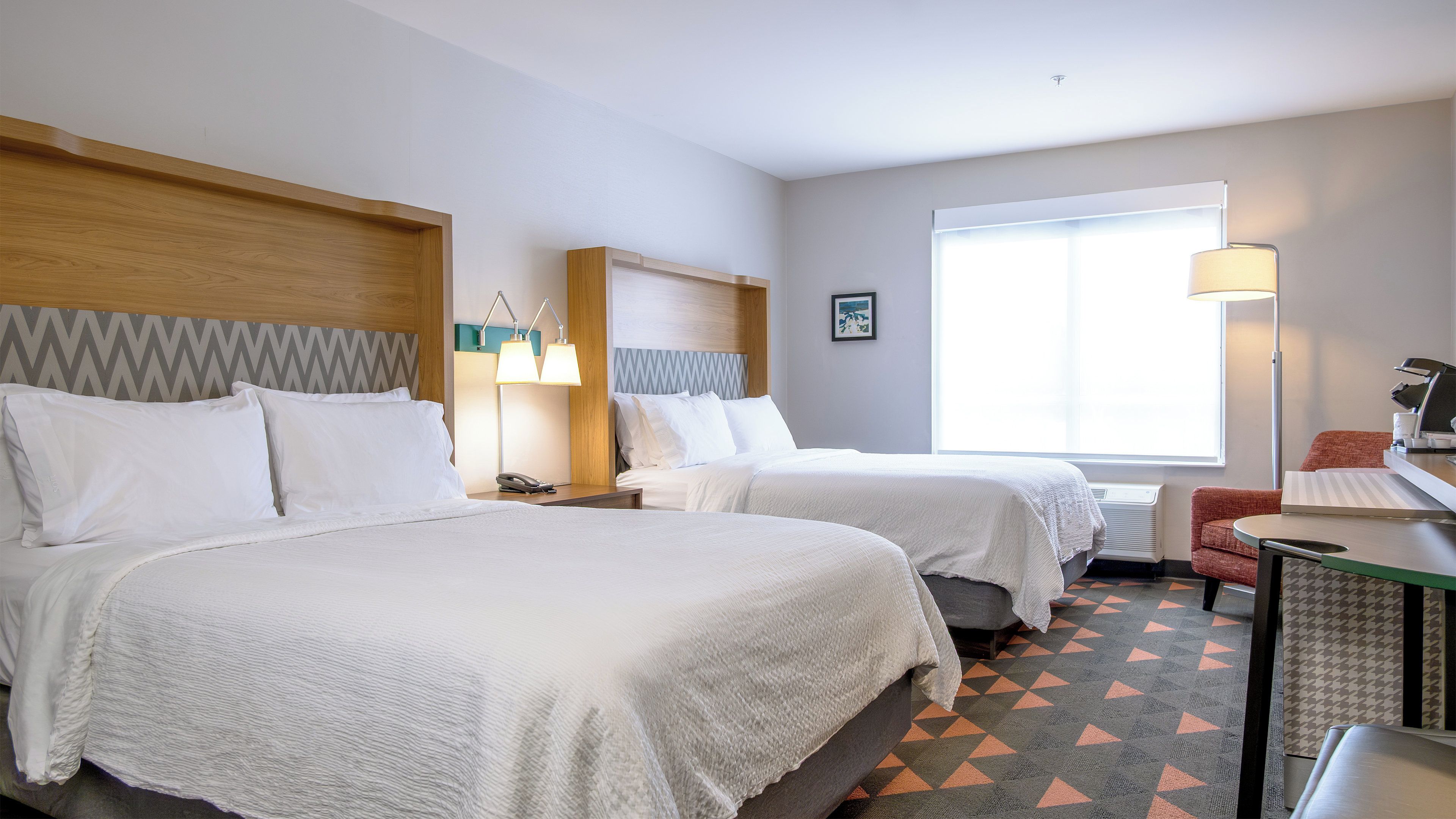 A hotel room with two beds, a desk, a chair, a window with blinds, and a wall-mounted lamp.
