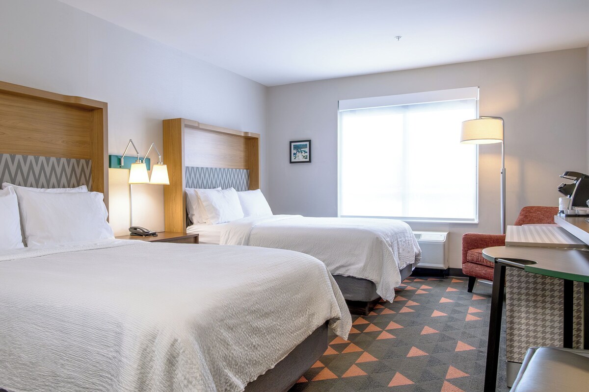A hotel room with two beds, a desk, a chair, a window with blinds, and a wall-mounted lamp.