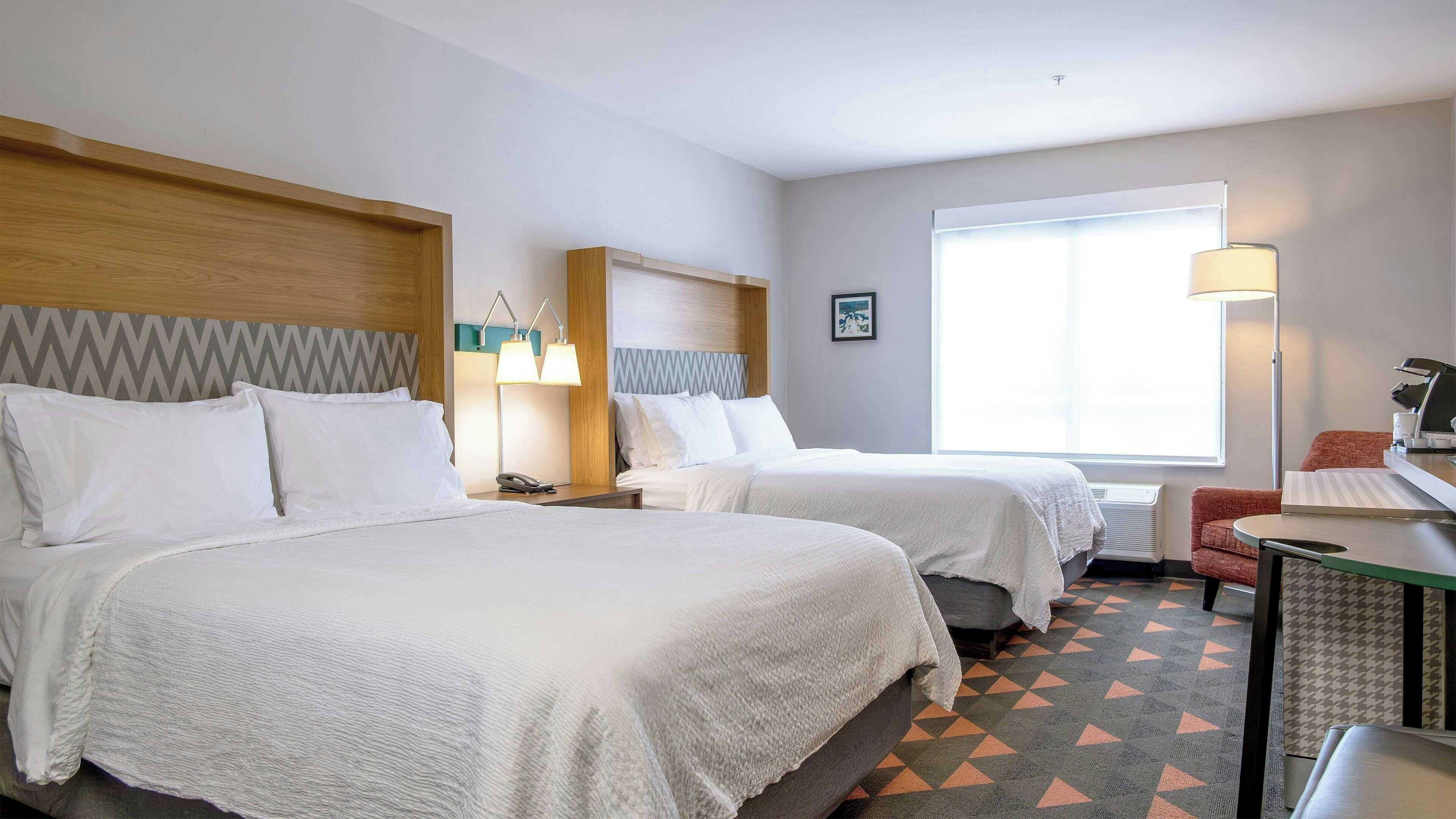 A hotel room with two beds, a desk, a chair, a window with blinds, and a wall-mounted lamp.