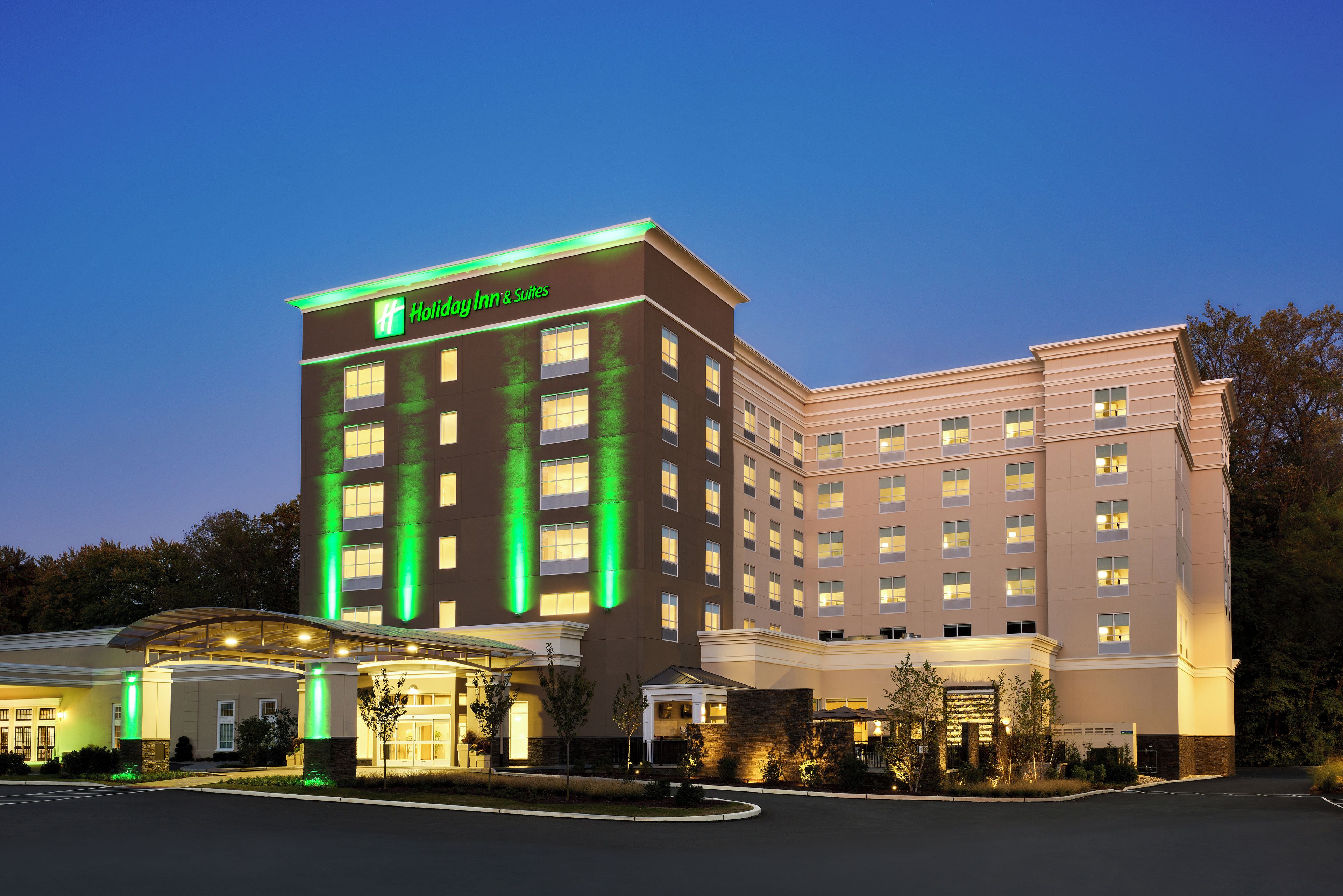 Un hotel Holiday Inn & Suites con diseño moderno, que cuenta con un llamativo letrero verde, varios pisos y una entrada bien iluminada.