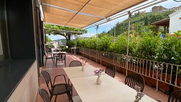Un patio exterior con mesas y sillas, un toldo y vistas a una colina.