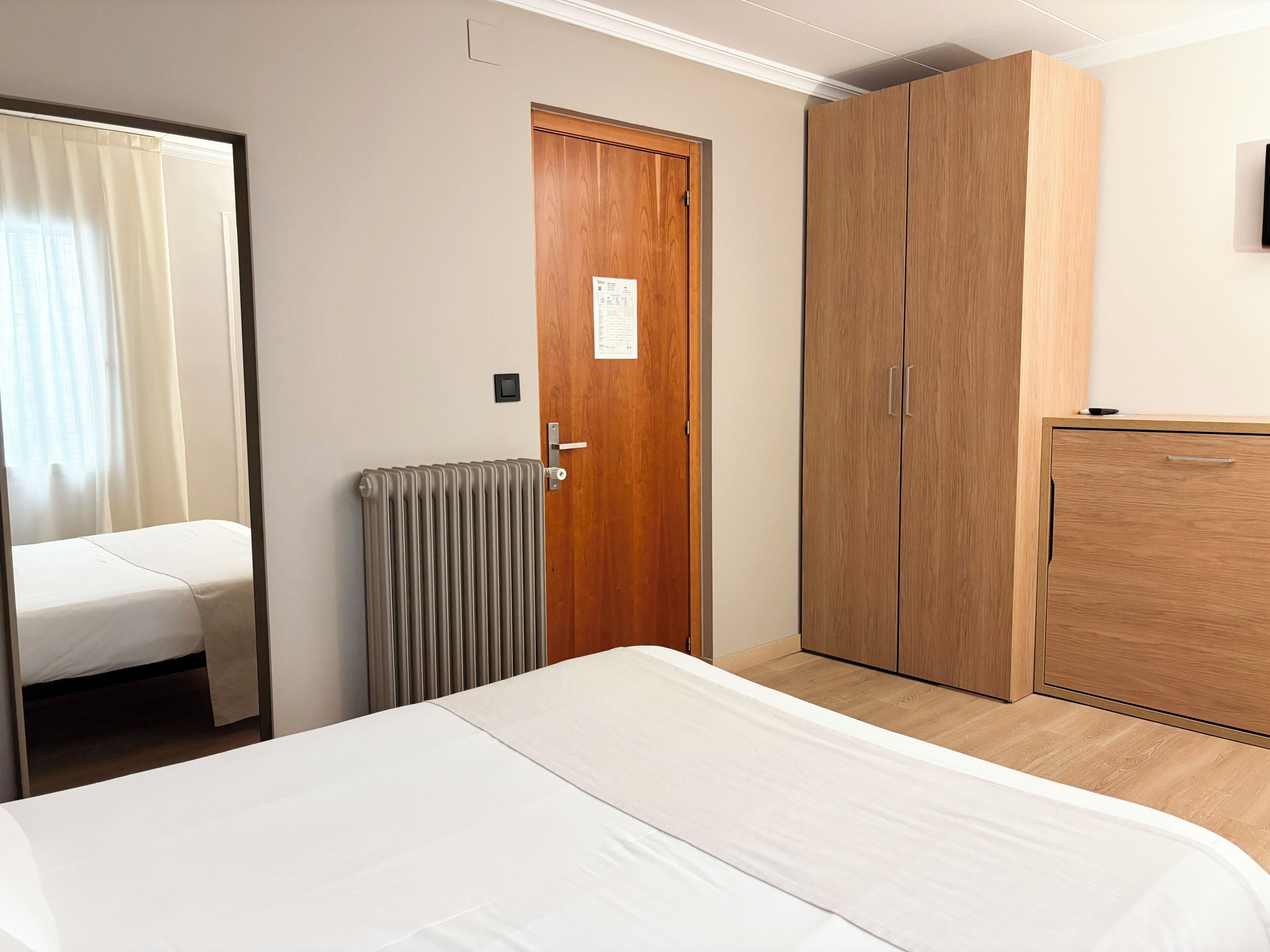 Habitación de hotel con cama, armario, radiador y puerta con un cartel.