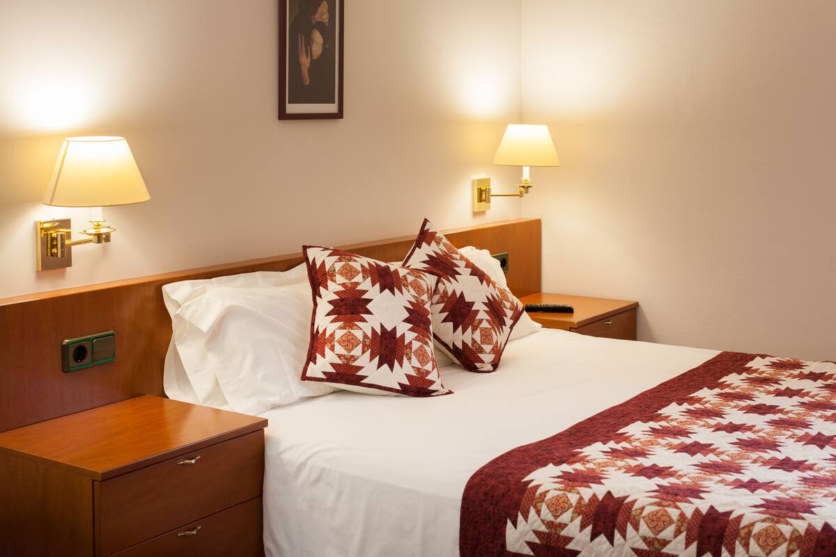 Ein Hotelzimmer mit einem ordentlich bezogenem Bett, zwei Nachttischlampen und einem Nachttisch mit Schubladen.