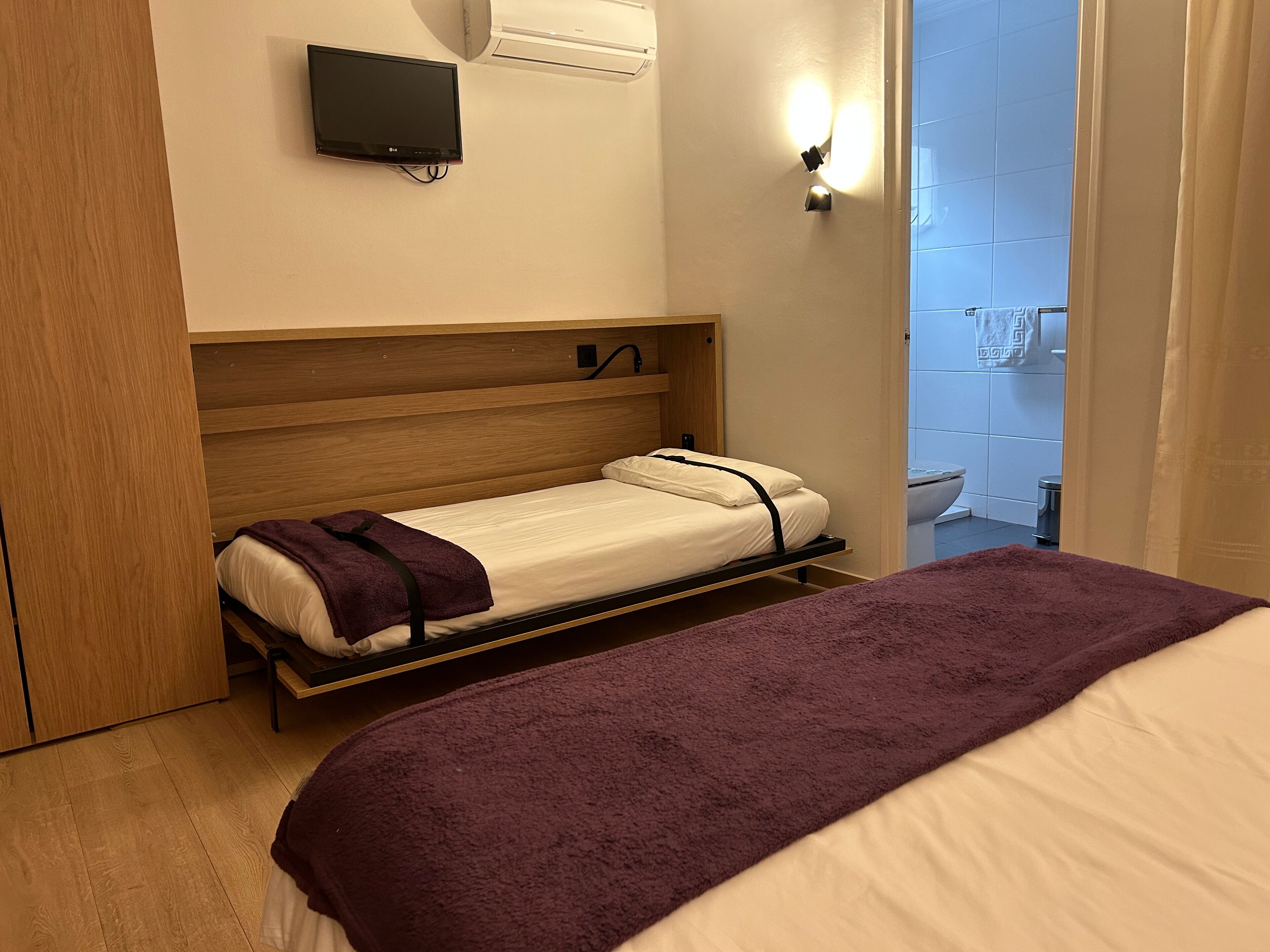 Una habitación de hotel compacta con una cama individual, cabecera de madera, televisor de pantalla plana montado en la pared y lámpara fijada a la pared.