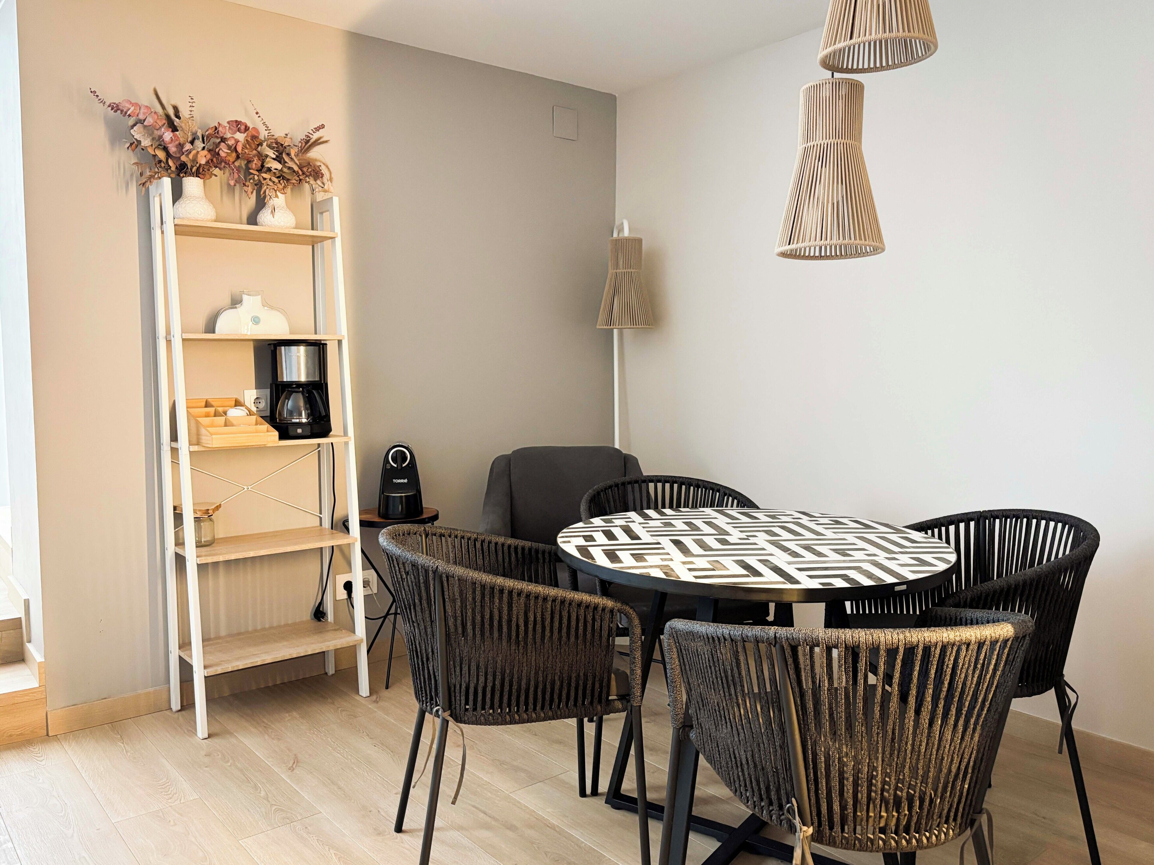A dining area with a round table, four chairs, a coffee machine, and a shelf with decorative items.