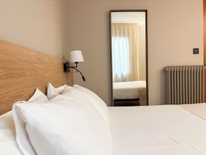 Habitación de hotel con cabecera de madera, cama con ropa blanca, lámpara de pared, espejo que refleja una ventana con cortinas y un radiador.