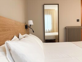 Habitación de hotel con cabecera de madera, cama con ropa blanca, lámpara de pared, espejo que refleja una ventana con cortinas y un radiador.