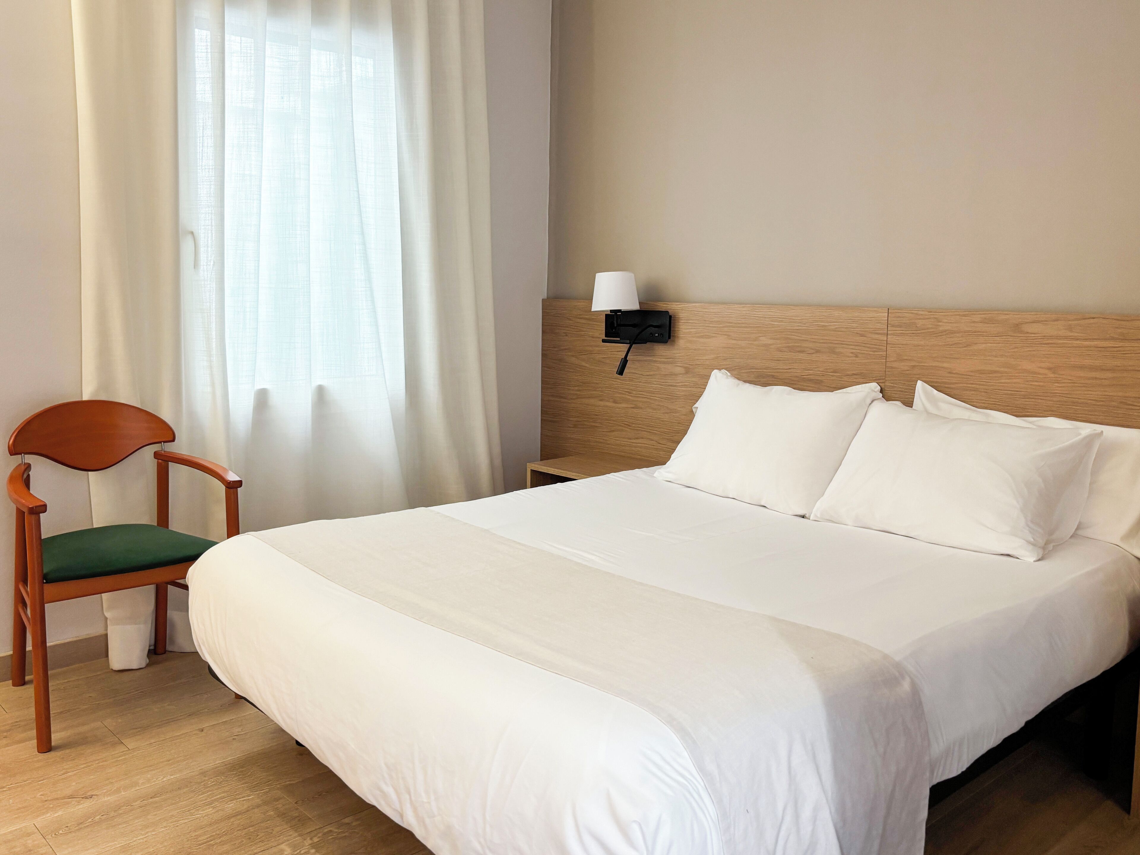 Una habitación de hotel con cabecera de madera, una cama individual con ropa de cama blanca, una silla de madera con cojín verde y una mesita de noche con una lámpara.