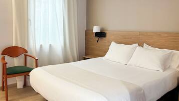 Una habitación de hotel con cabecera de madera, una cama individual con ropa de cama blanca, una silla de madera con cojín verde y una mesita de noche con una lámpara.
