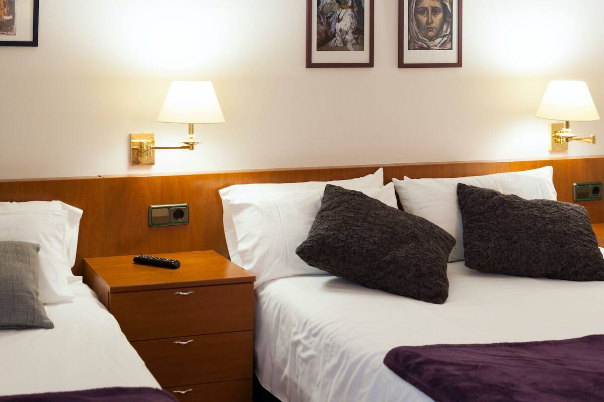 Ein Hotelzimmer mit zwei Betten, jeweils mit weißen Leinen und dunklen Kissen mit Muster. An den Betten befinden sich Nachttische mit Lampen, einer Fernbedienung und einer Schublade. Das Zimmer hat ein Holz-Kopfteil und eine Wandleuchte.