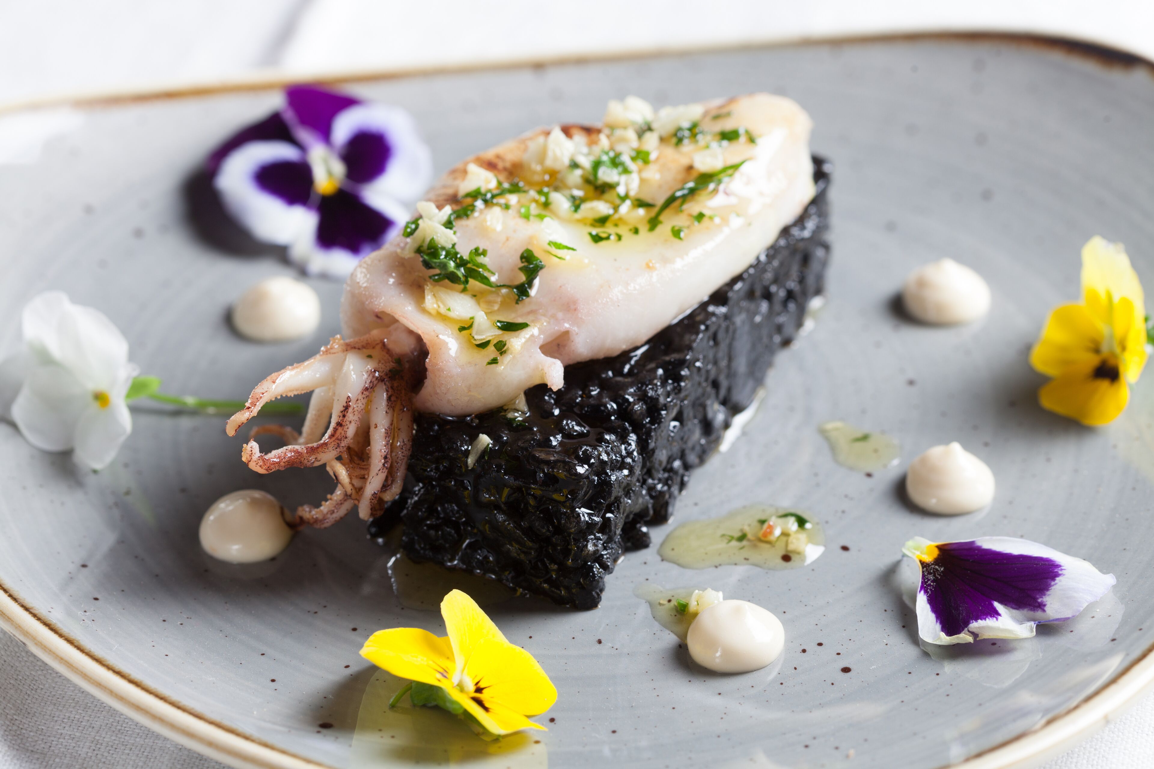 A seafood dish with squid, garnished with herbs, served on a bed of black seaweed, surrounded by edible flowers on a ceramic plate.