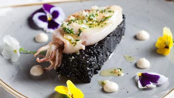 A seafood dish with squid, garnished with herbs, served on a bed of black seaweed, surrounded by edible flowers on a ceramic plate.