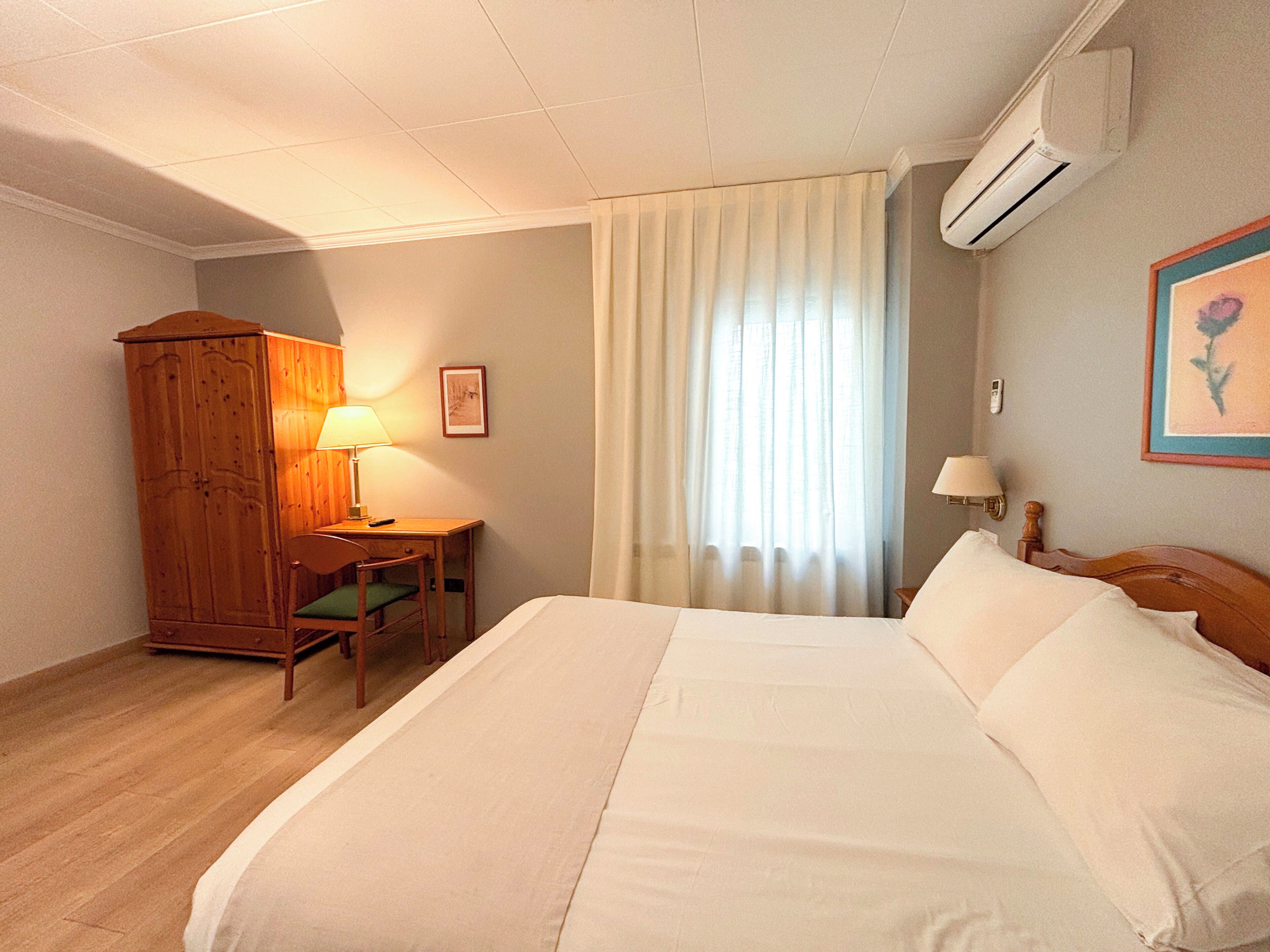 A hotel room with a large bed, a wooden wardrobe, a desk with a chair, a window with curtains, and an air conditioning unit.
