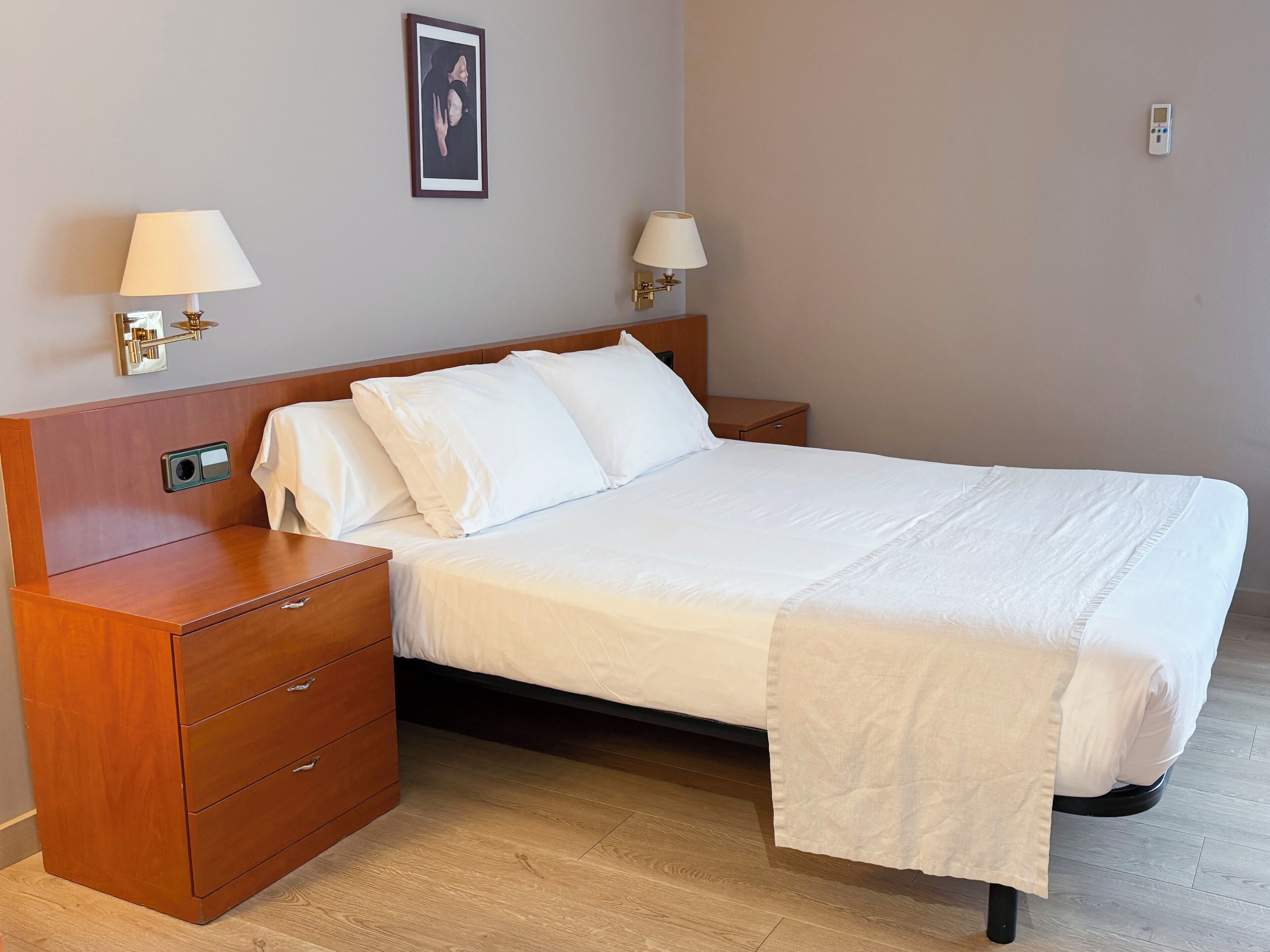 A neatly made bed with white linens, a wooden nightstand with a lamp, and a wall-mounted picture.