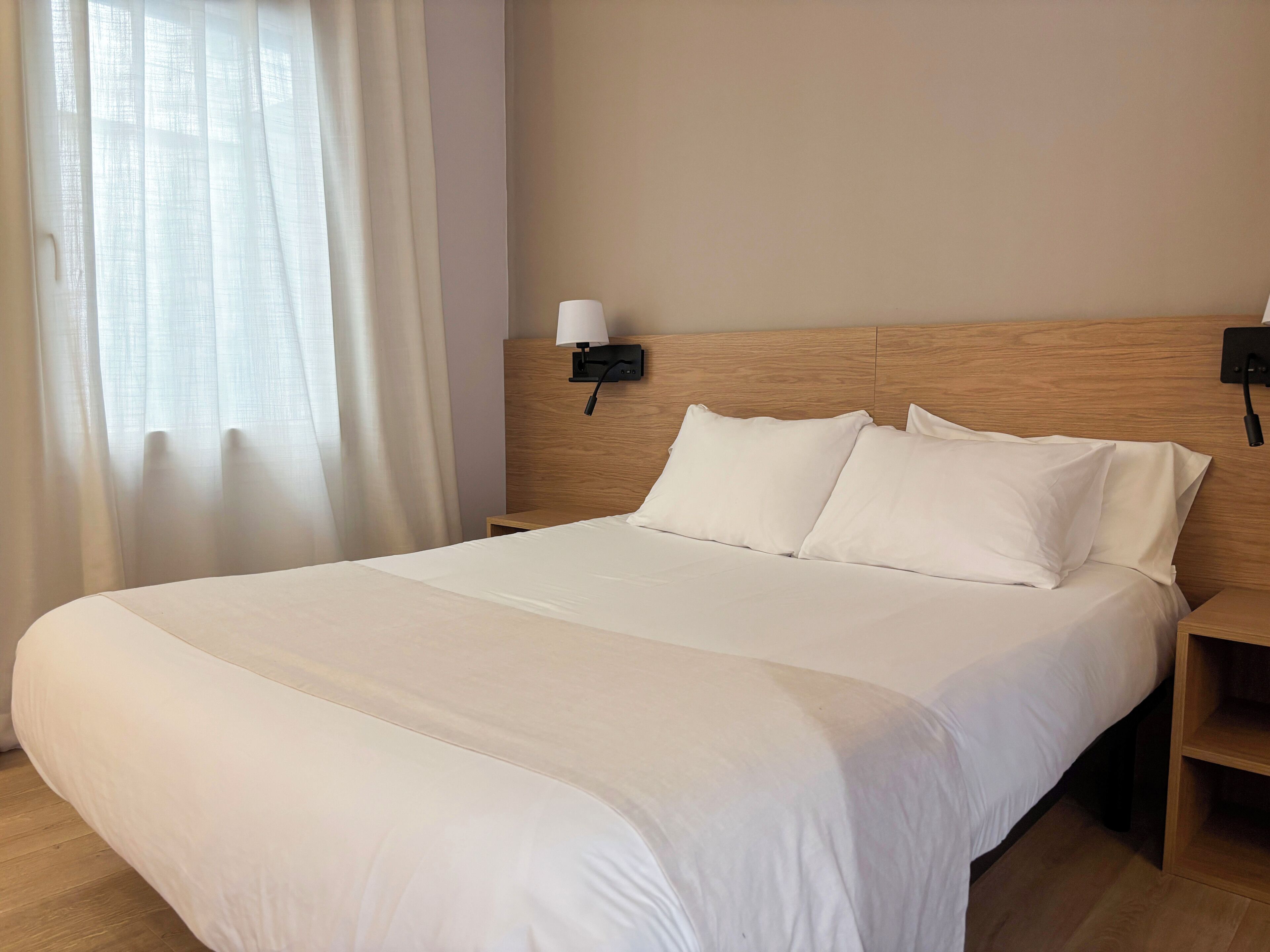 Una habitación de hotel con una cama grande, una mesita de noche, una lámpara de pared y cortinas traslúcidas.