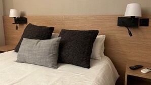 Una cama bien hecha con ropa de cama blanca y gris, dos almohadas negras con textura y un cabecero de madera.