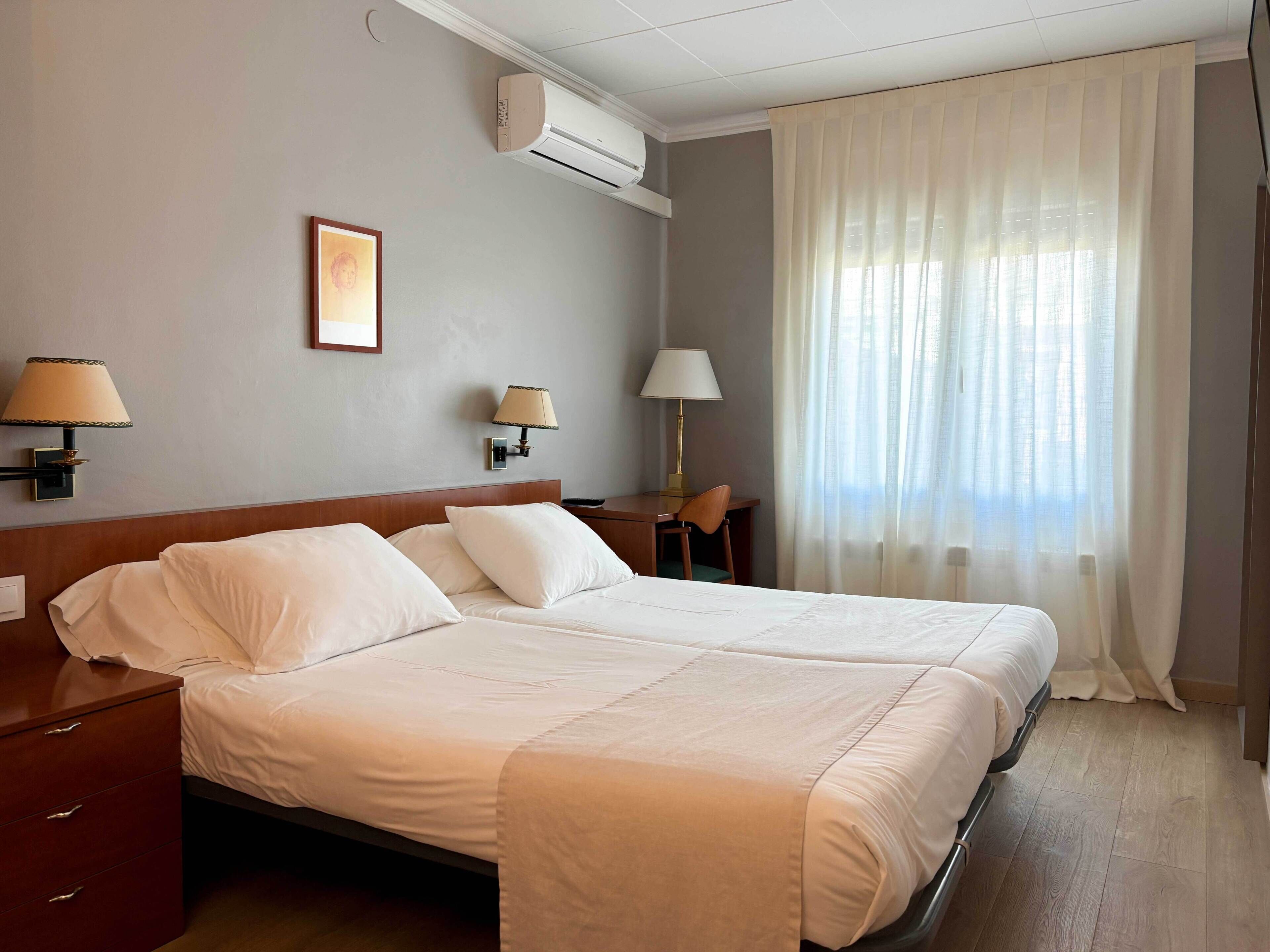 Habitación de hotel con dos camas, un escritorio, una silla, una lámpara, aire acondicionado y una ventana con cortinas.
