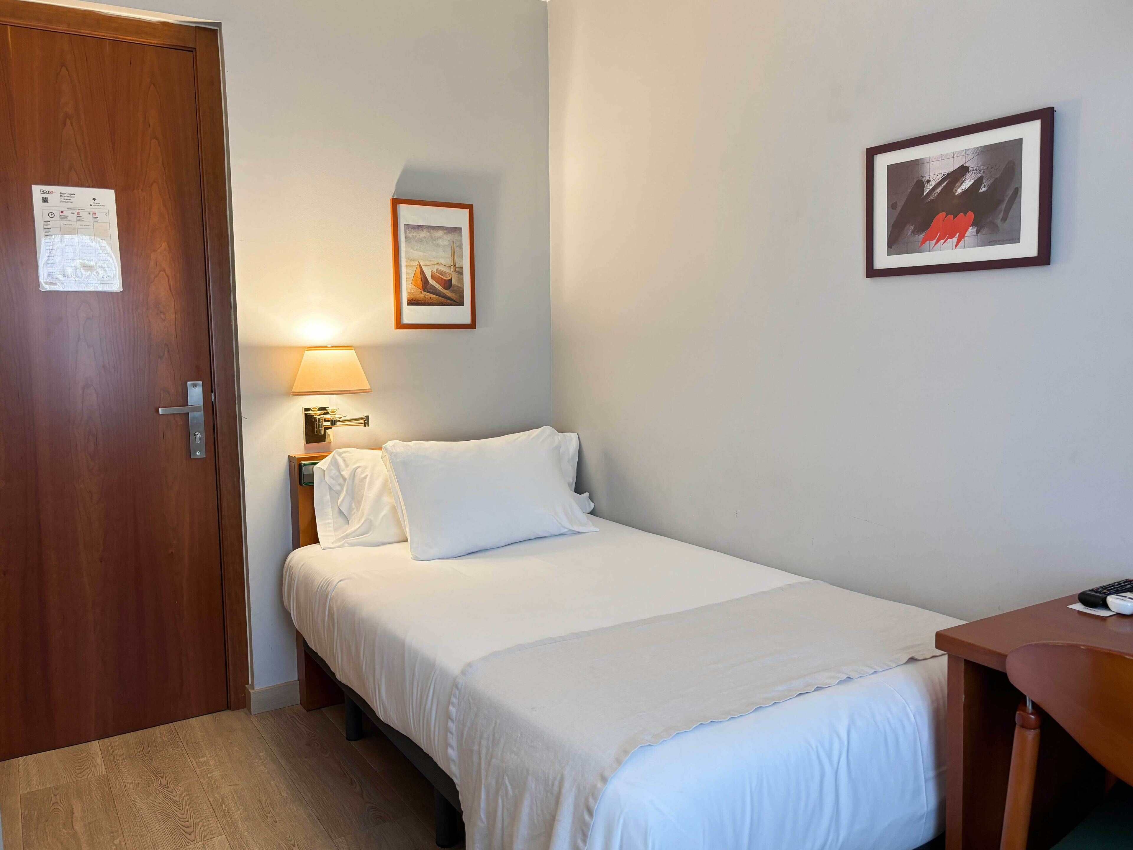 Habitación de hotel con puerta de madera, cama con ropa de cama blanca, mesita de noche con lámpara y un cuadro enmarcado en la pared.