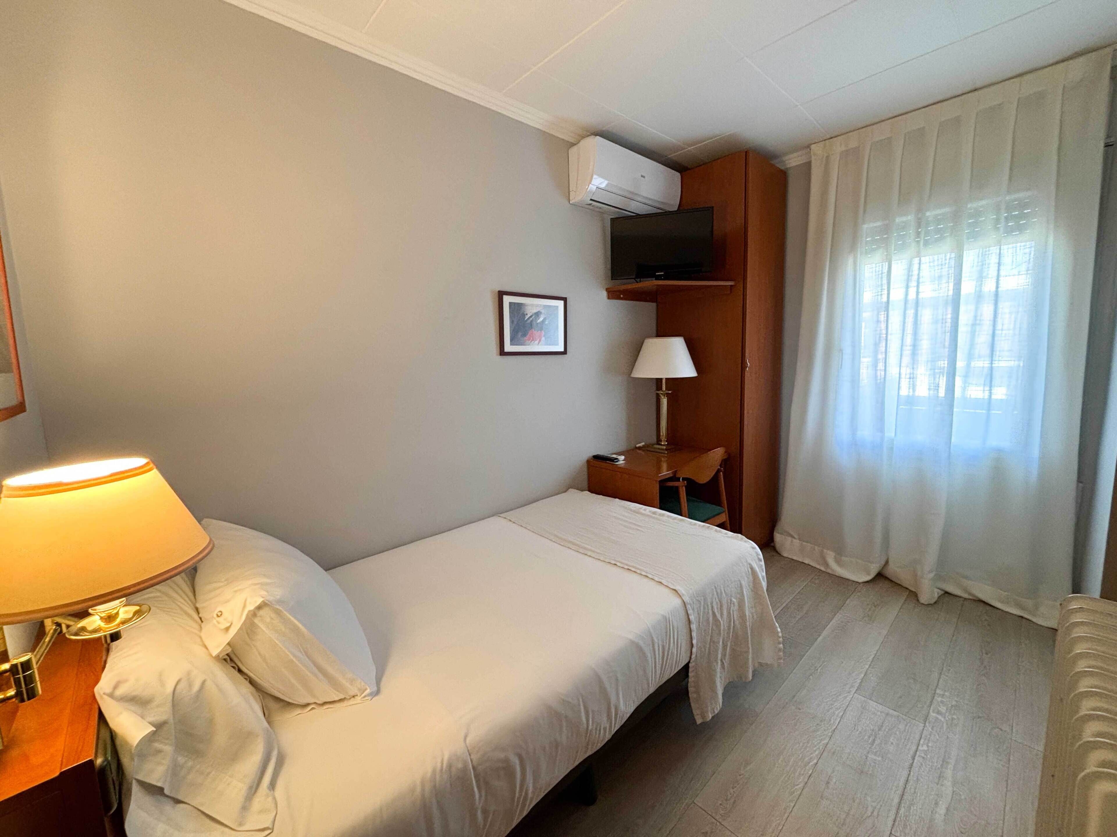 A hotel room with a bed, a desk with a lamp, a television, and a window with curtains.