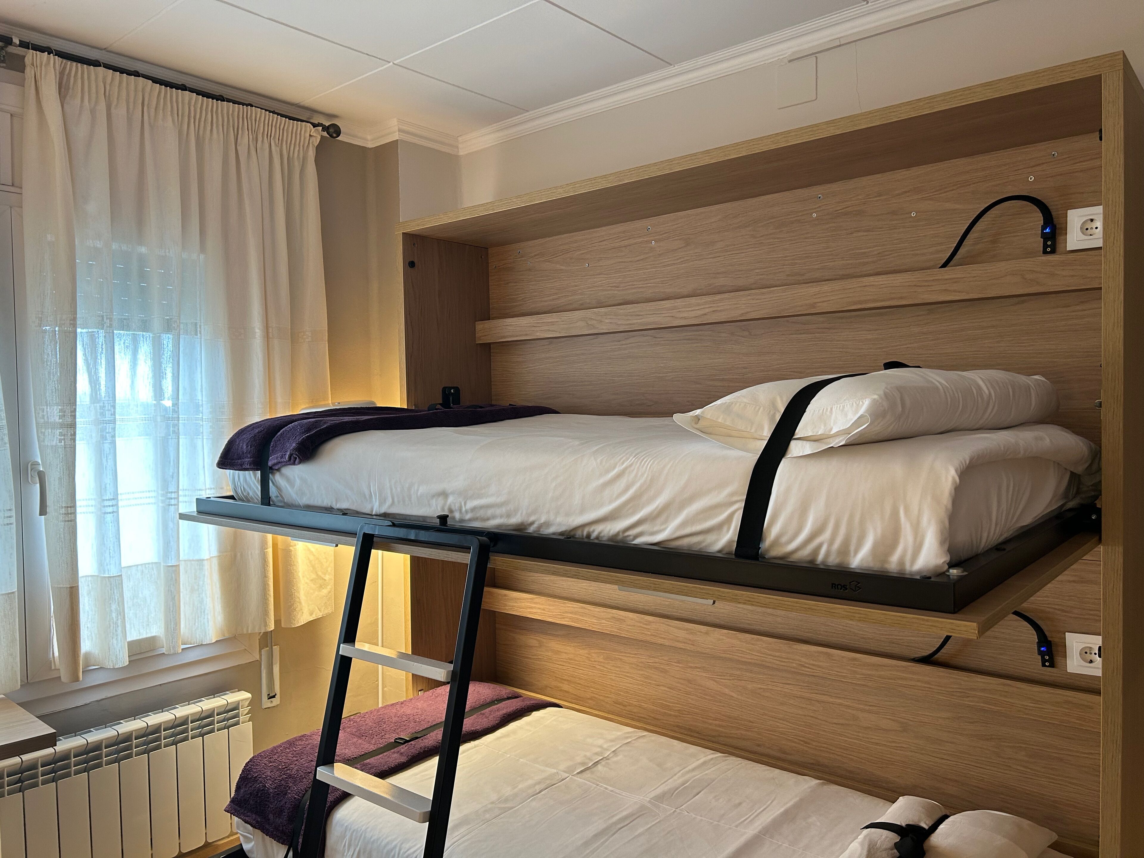 A bunk bed with a ladder, a window with curtains, and a radiator.