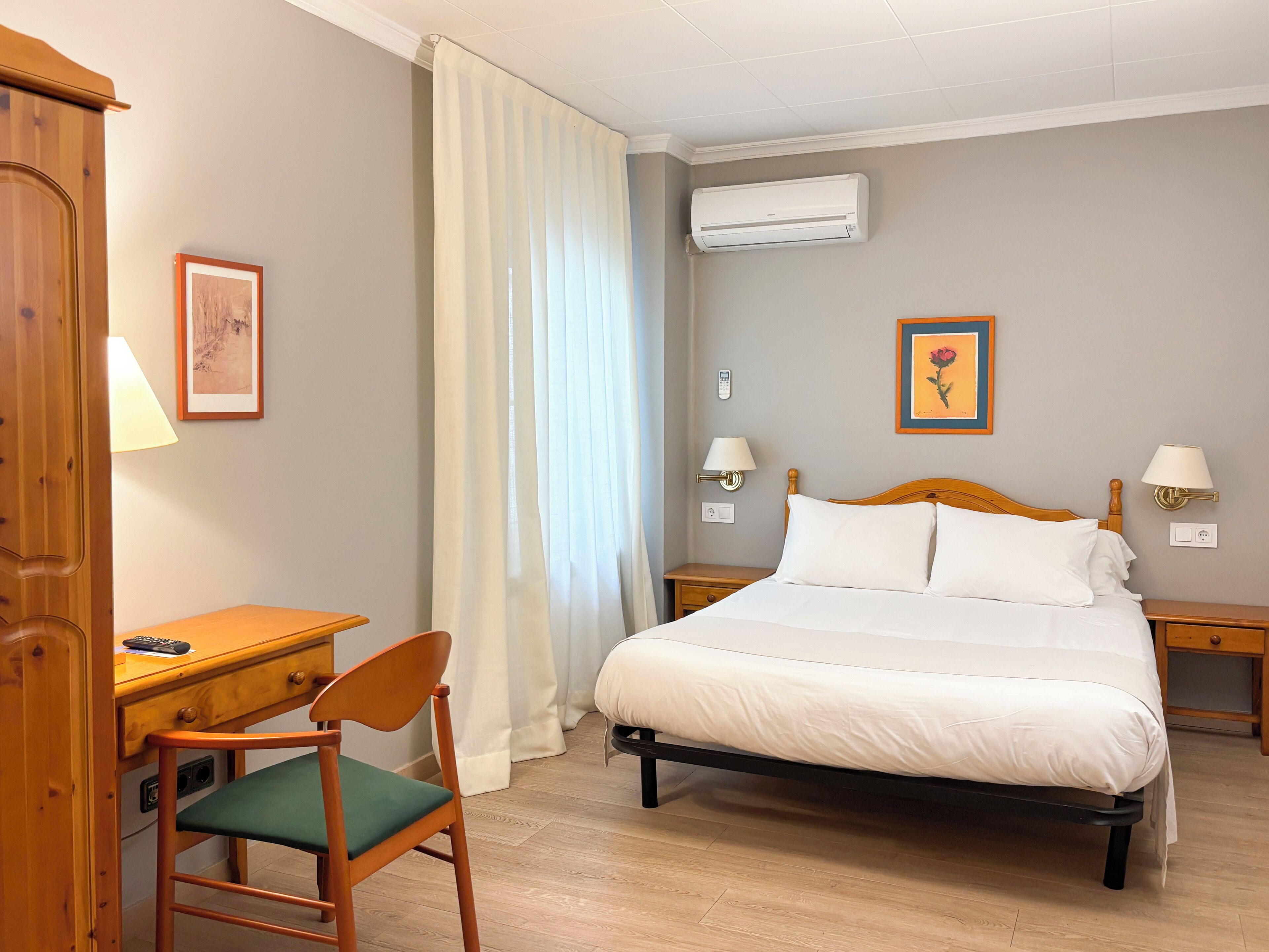 A hotel room with a bed, bedside tables, a desk, a chair, an air conditioner, and two wall-mounted lamps.