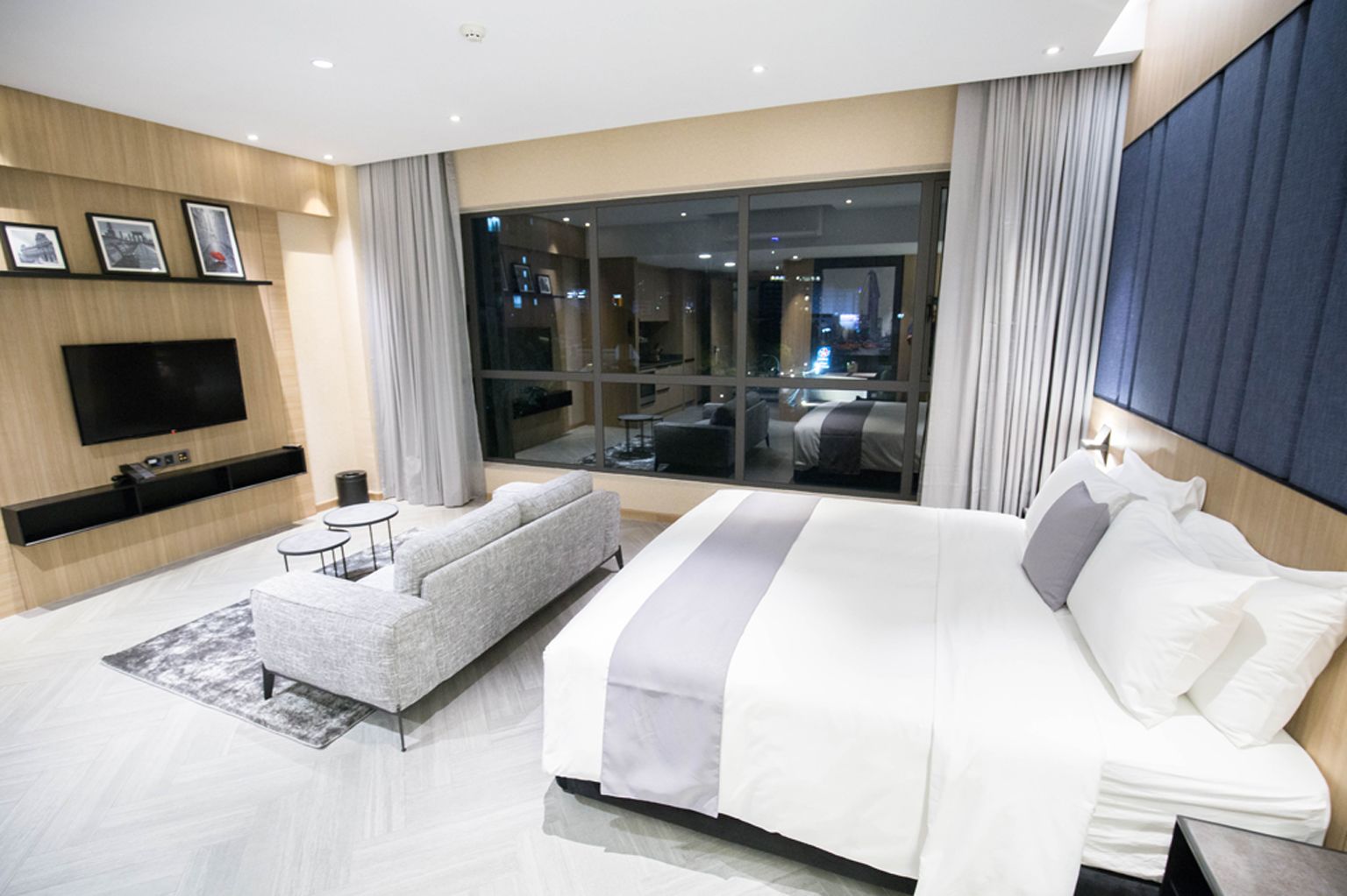 A modern hotel room with a large bed, a sofa, a flat-screen TV, and a large window offering a city view.
