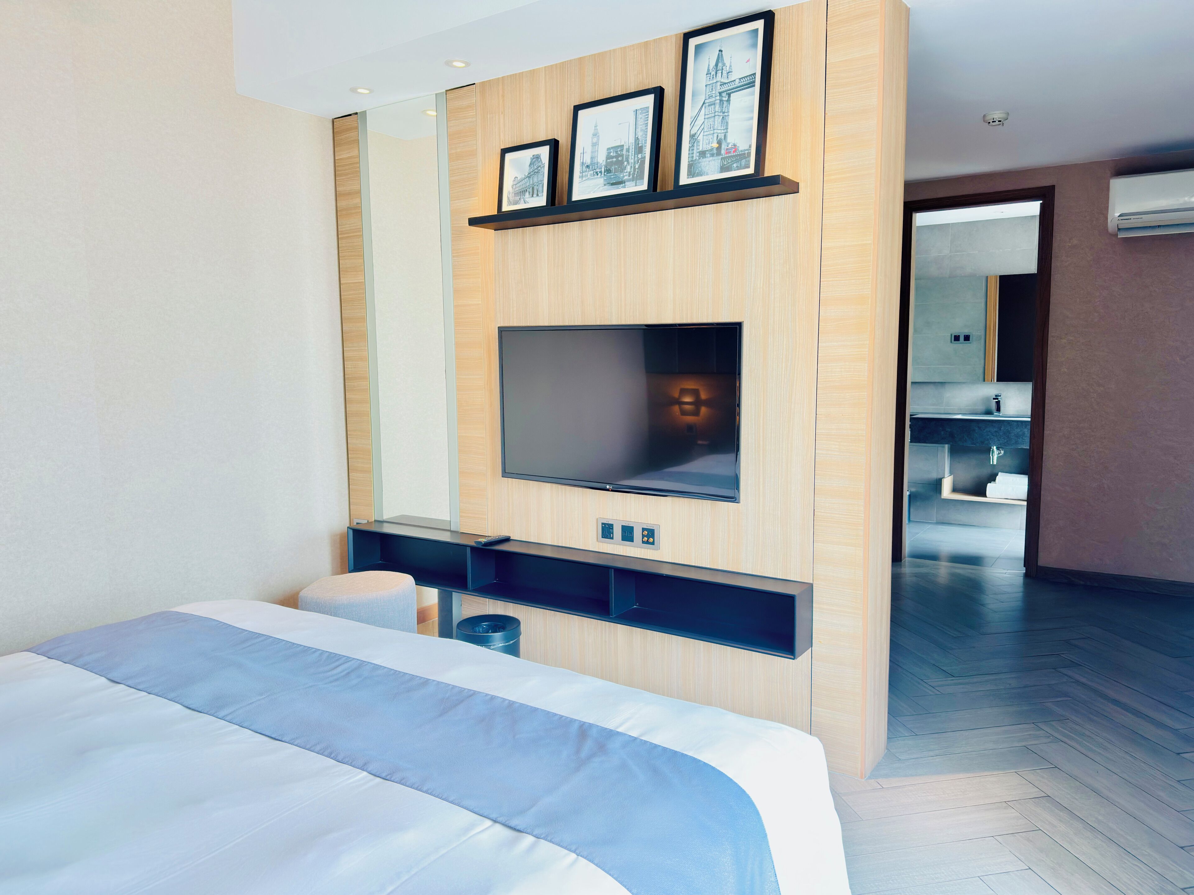 Ett modernt hotellrum med en stor säng, en platt-TV som är monterad på en träpanel, ett litet skrivbord med en stol och ett badrum som syns genom en öppen dörr.