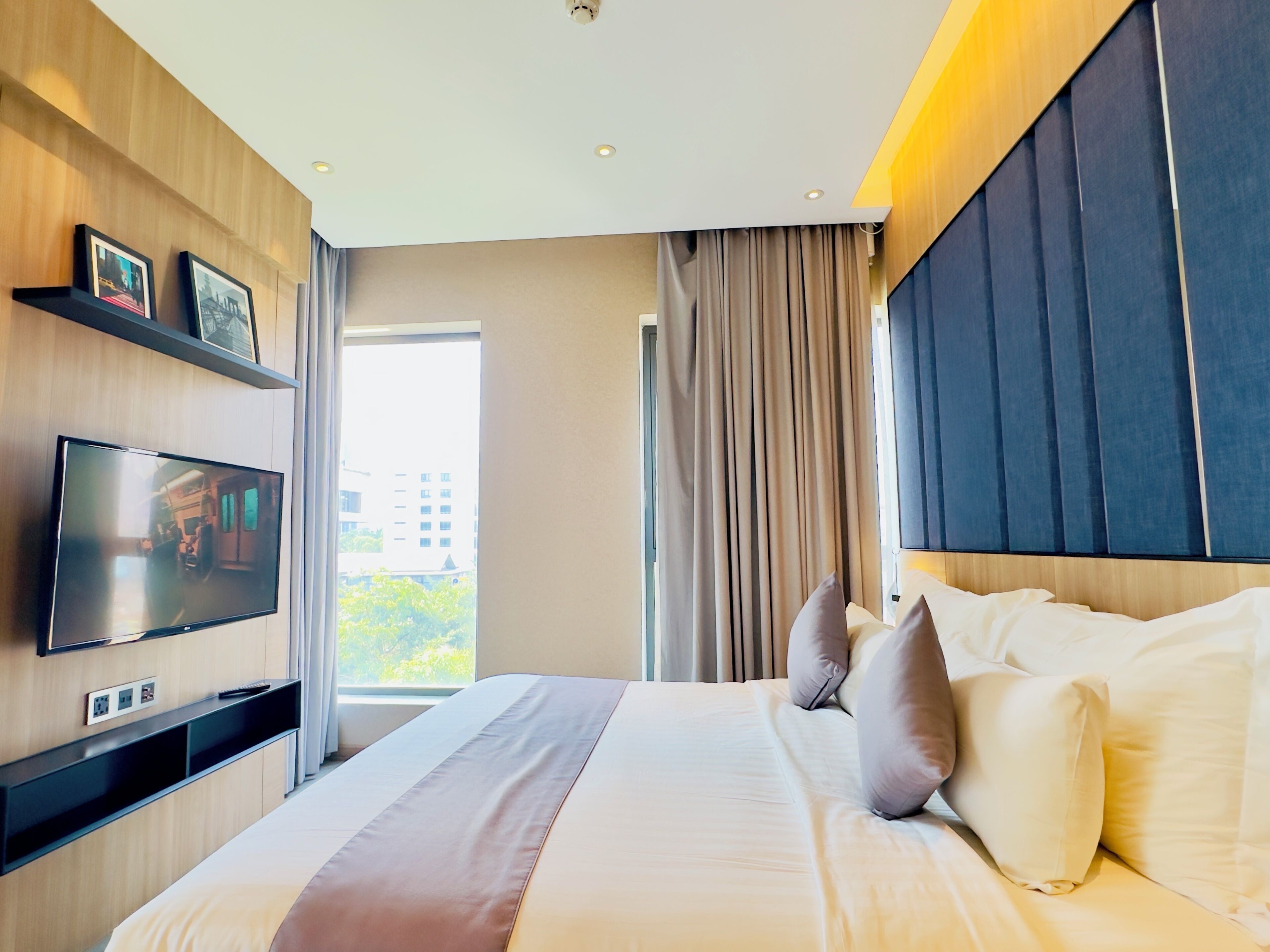 A modern hotel room with a large bed, a flat-screen TV mounted on the wall, a window with curtains, and a city view.