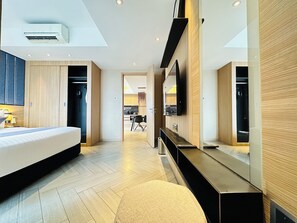 Quarto de hotel moderno com uma cama, toucador com espelho, televisão de tela plana e uma kitchenette ao fundo.