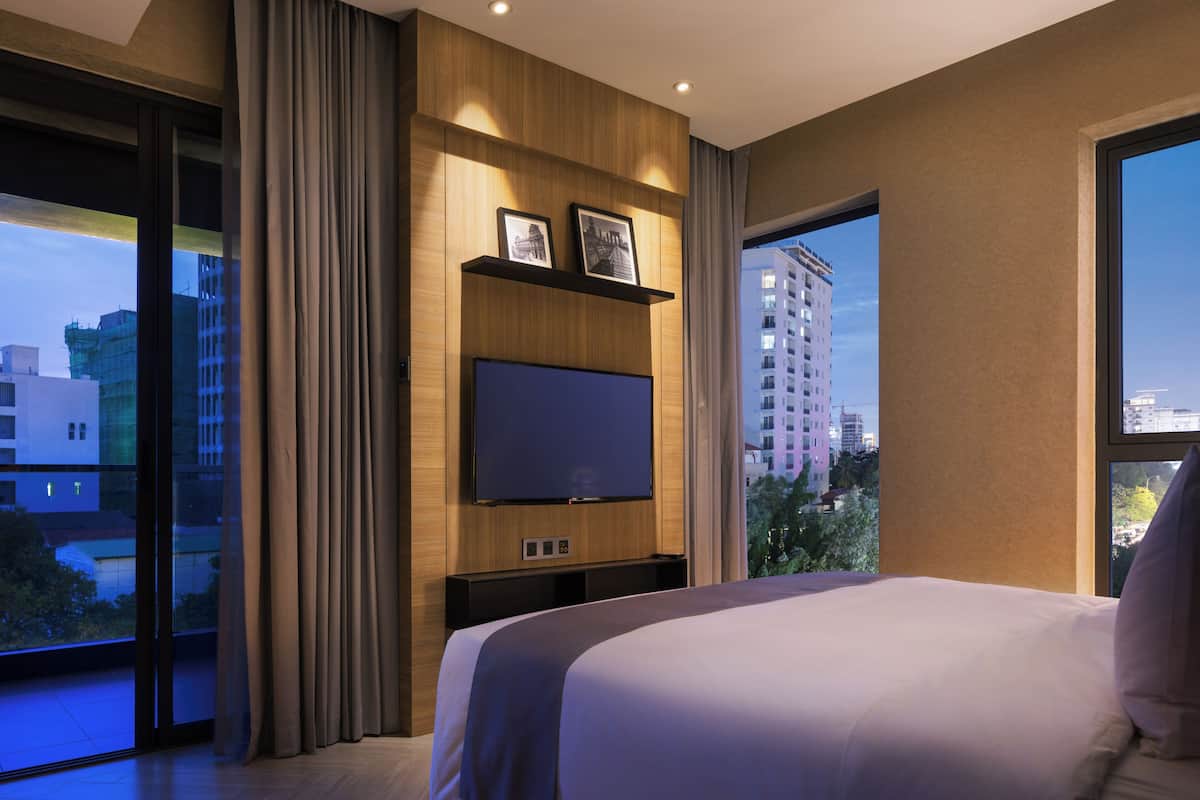 Une chambre d’hôtel moderne dotée d’un grand lit, d’un téléviseur à écran plat fixé sur un panneau en bois, d’une porte-fenêtre coulissante donnant sur un balcon et offrant une vue sur la ville.