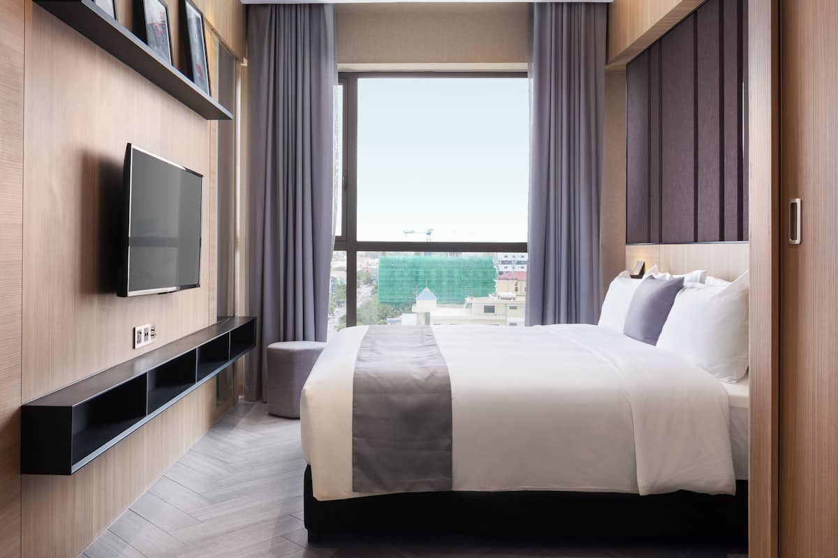 Une chambre d’hôtel moderne avec un grand lit, un téléviseur à écran plat fixé au mur et une vue sur le paysage urbain par la fenêtre.