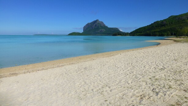 Een strand met helder turquoise water, een zandstrand en een opvallende rotsberg op de achtergrond.