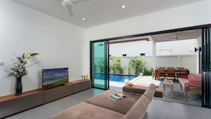 A modern living room with a large window, a flat-screen TV, a sofa, and a dining area with wooden furniture. - Large 3BR Villa with Big Pool by Intira (Rawai)