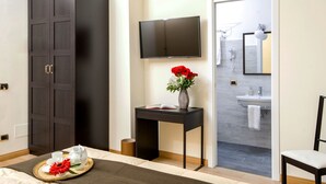 A hotel room with a dark wardrobe, a bedside table with a vase of red flowers, a flat-screen TV mounted on the wall, and a view into the bathroom. - Mybedcavour (Rome)