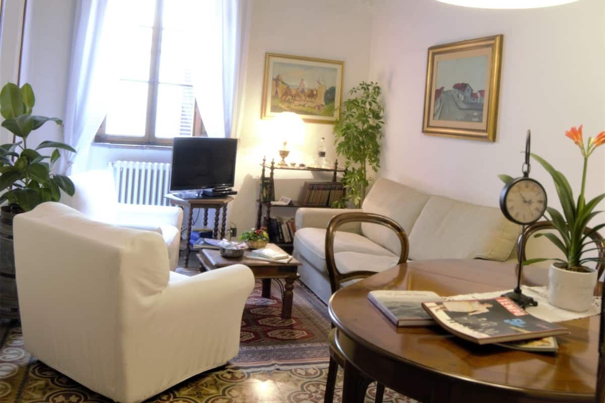 Ein Wohnzimmer mit einem weißen Sofa, einem Holztisch und einem Fernseher. Der Boden weist ein Muster auf und die Decke hat sichtbare Balken.