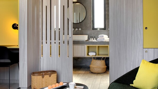 Une salle de bain moderne aux couleurs gris et jaune, avec un miroir rond, un lavabo et une petite fenêtre.