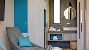 Une chambre moderne avec un mur d’accent bleu, un miroir rond, une petite table ronde avec des verres et un coin salon agrémenté d’un coussin bleu.