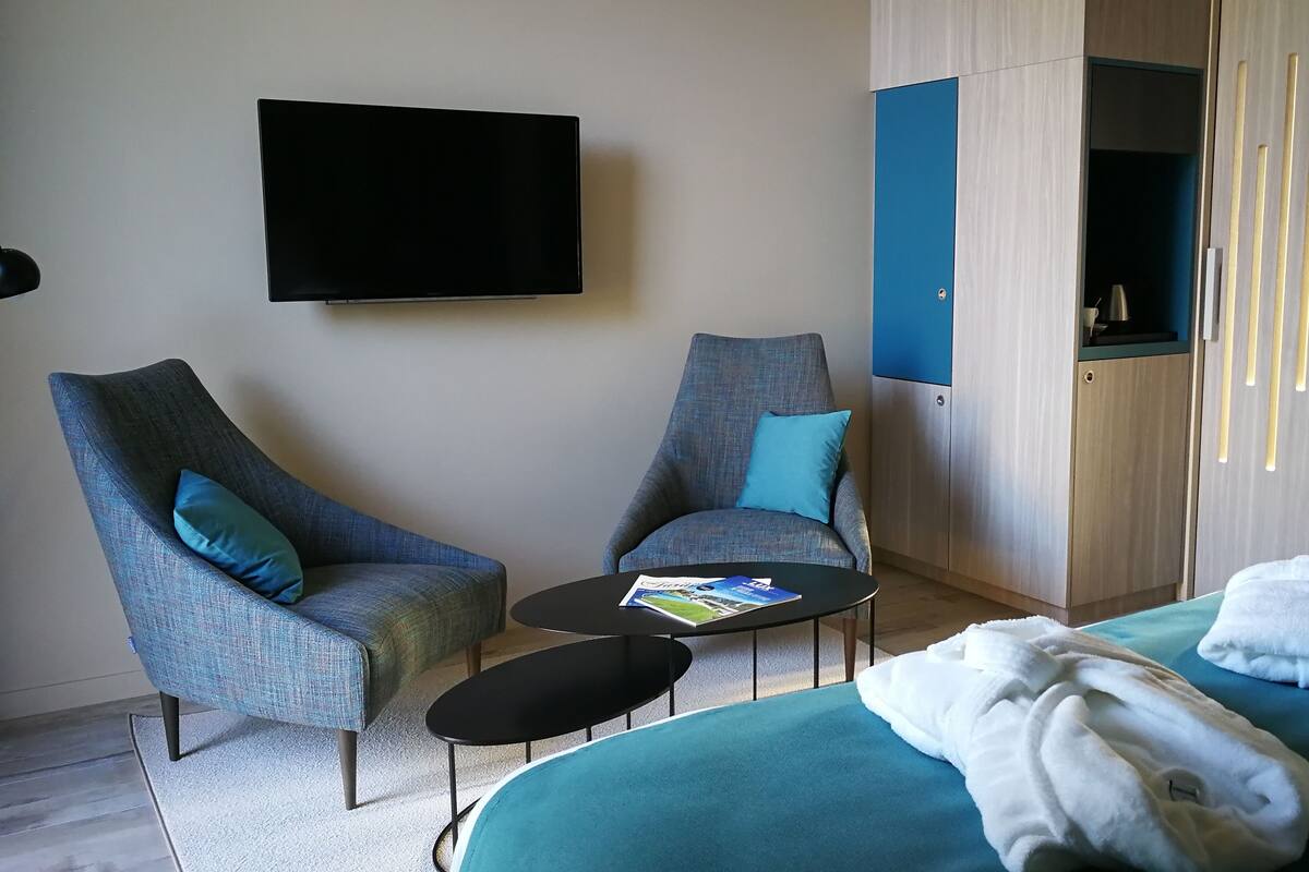 Una habitación de hotel moderna con dos sillones, una mesita, una cama con toallas, un televisor y un armario.