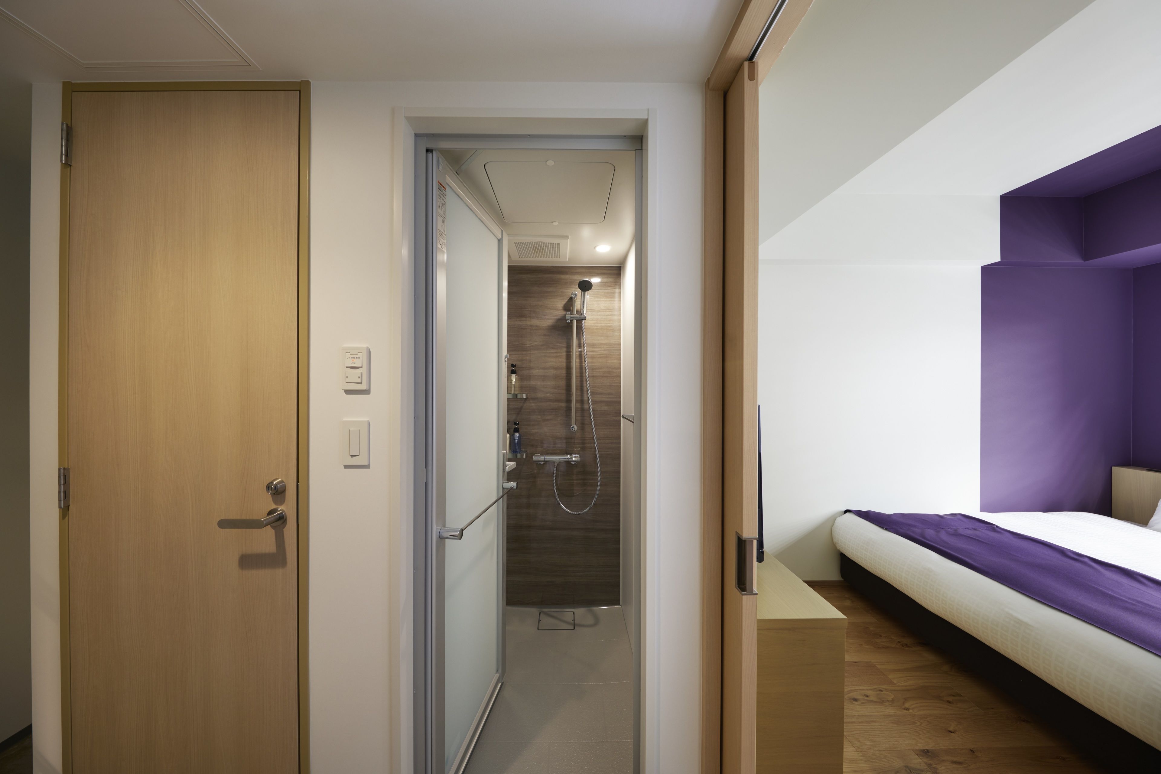 Una habitación de hotel moderna con puerta de madera, zona de ducha, cama con cabecero púrpura y una mesita de noche pequeña.