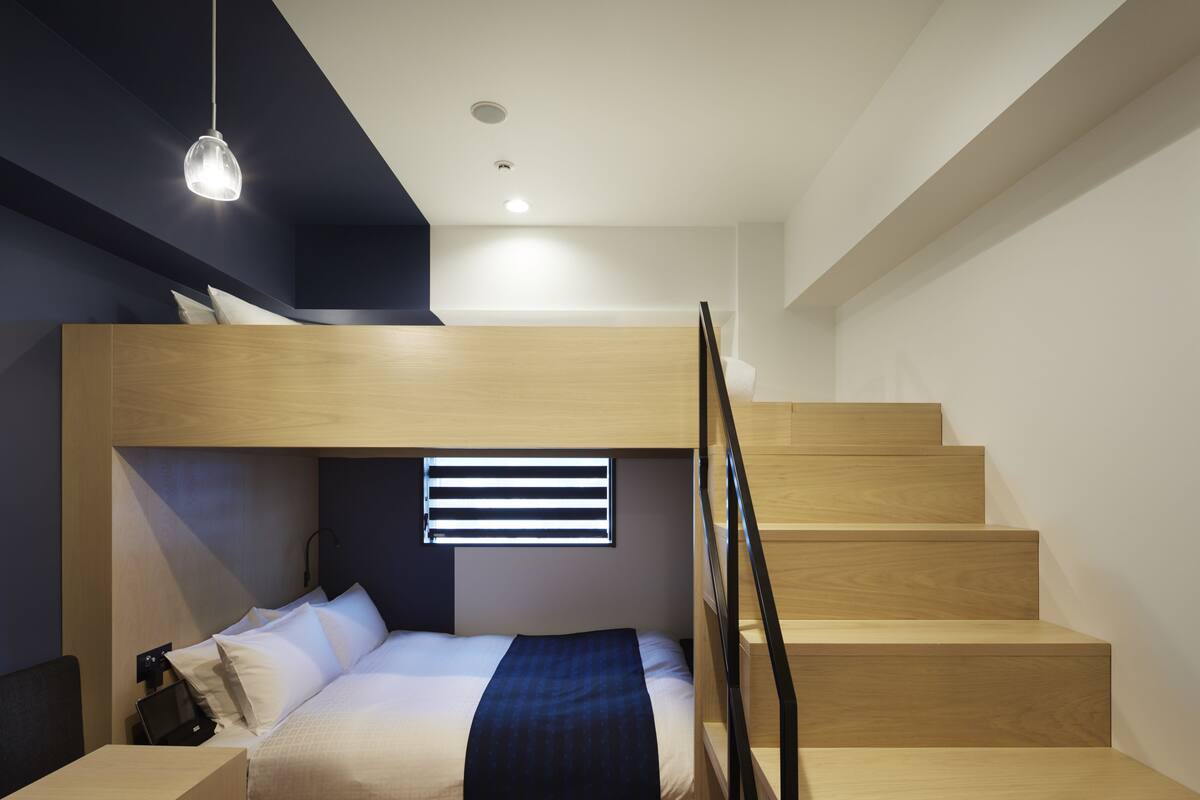 A modern bedroom with a bunk bed, wooden staircase, and a bedside table with a lamp.
