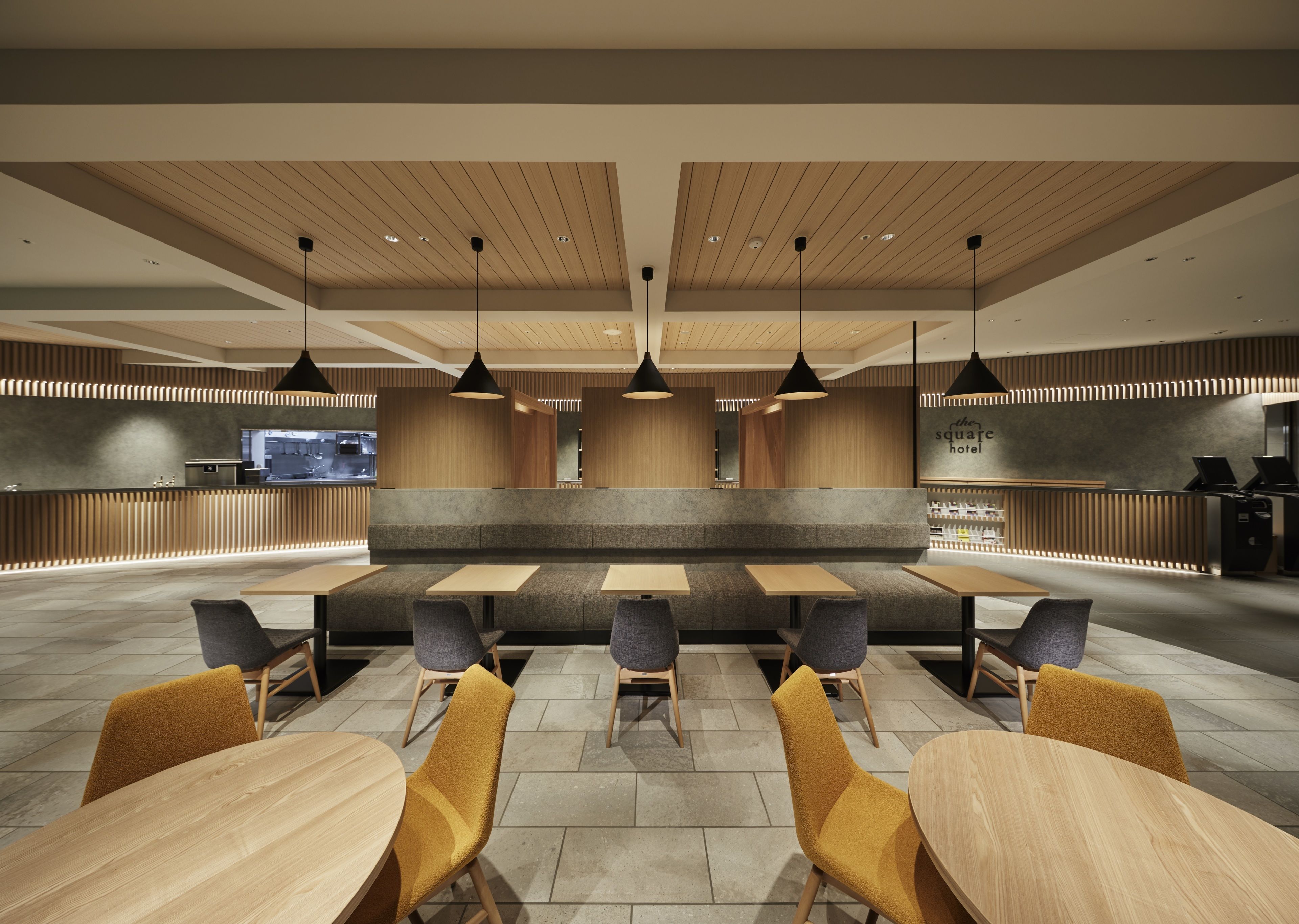 A modern restaurant with wooden ceiling panels, black pendant lights, and a central seating area with high stools and tables.