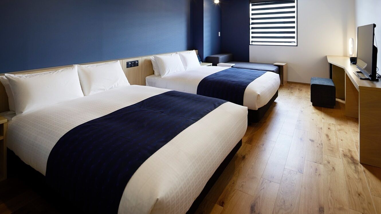 Une chambre d’hôtel avec deux lits, un bureau, une chaise, une télévision et une fenêtre avec des stores.