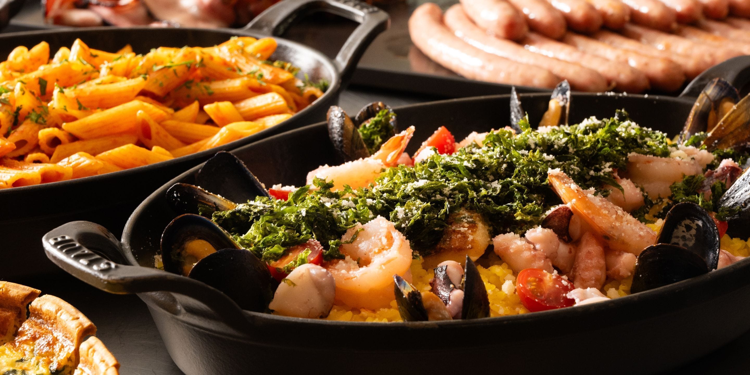 A variety of seafood dishes in a pan, including mussels, shrimp, and squid, with a side of pasta and sausages.