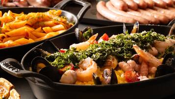 A variety of seafood dishes in a pan, including mussels, shrimp, and squid, with a side of pasta and sausages.