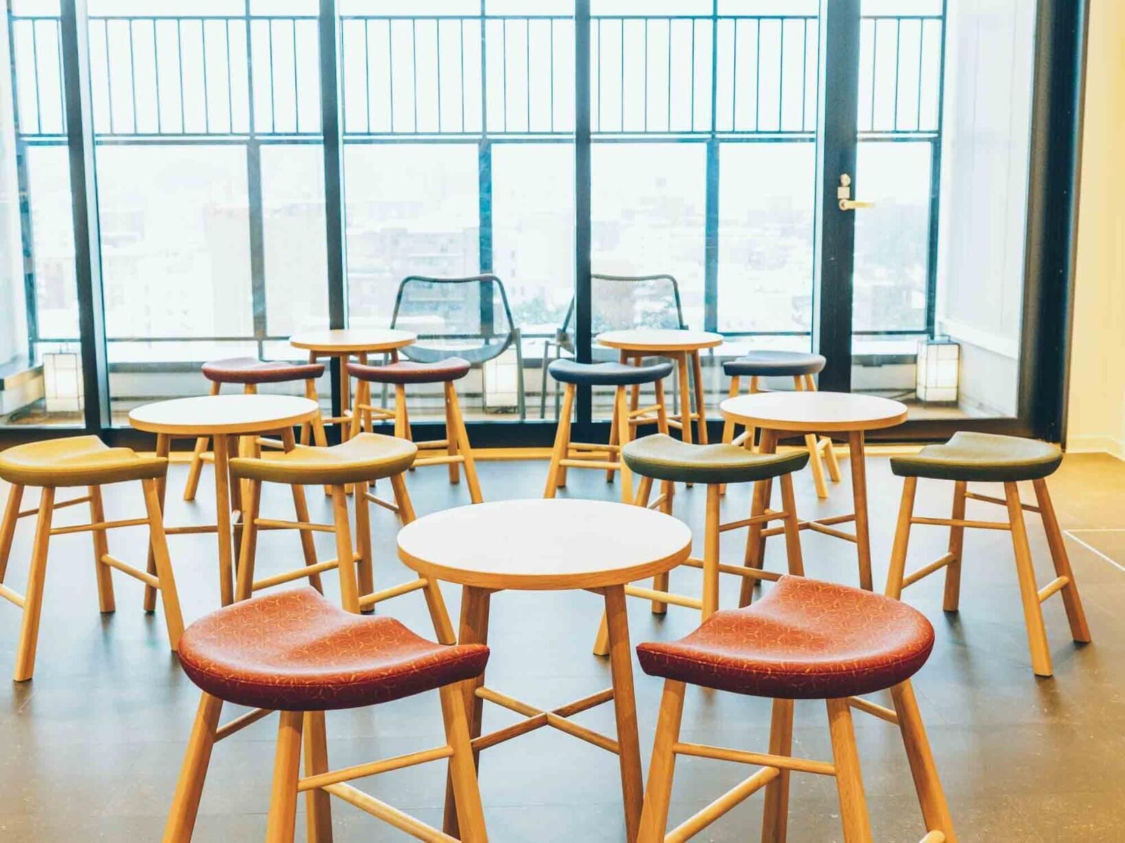 Un intérieur moderne avec des tables rondes et des tabourets disposés en cercle, de grandes fenêtres offrant une vue sur la ville et une porte dotée d’une poignée.