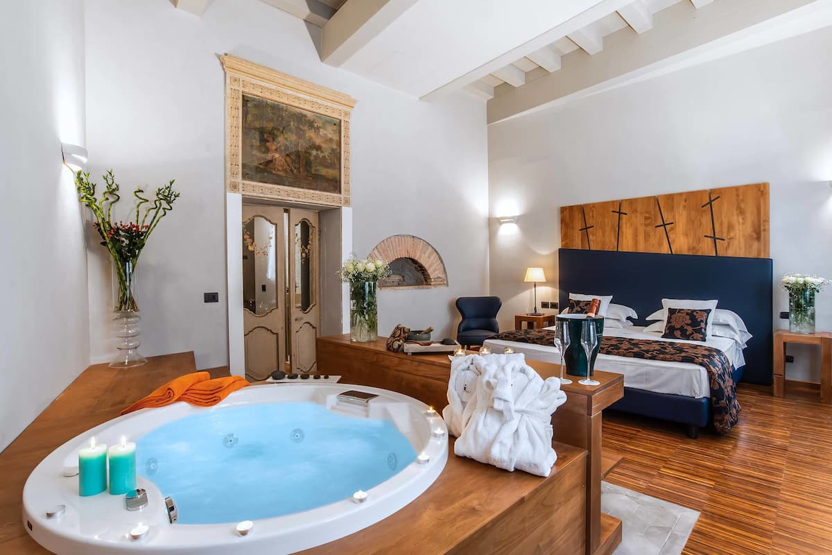 A modern hotel room with a large bathtub, a bed with a dark headboard, and a wooden floor.