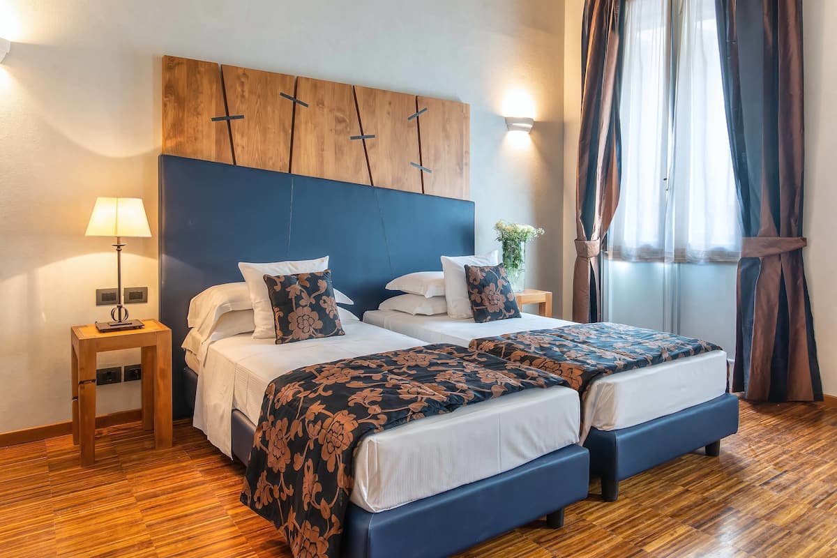 A hotel room with two beds, a wooden headboard, a bedside table with a lamp, and a window with curtains.
