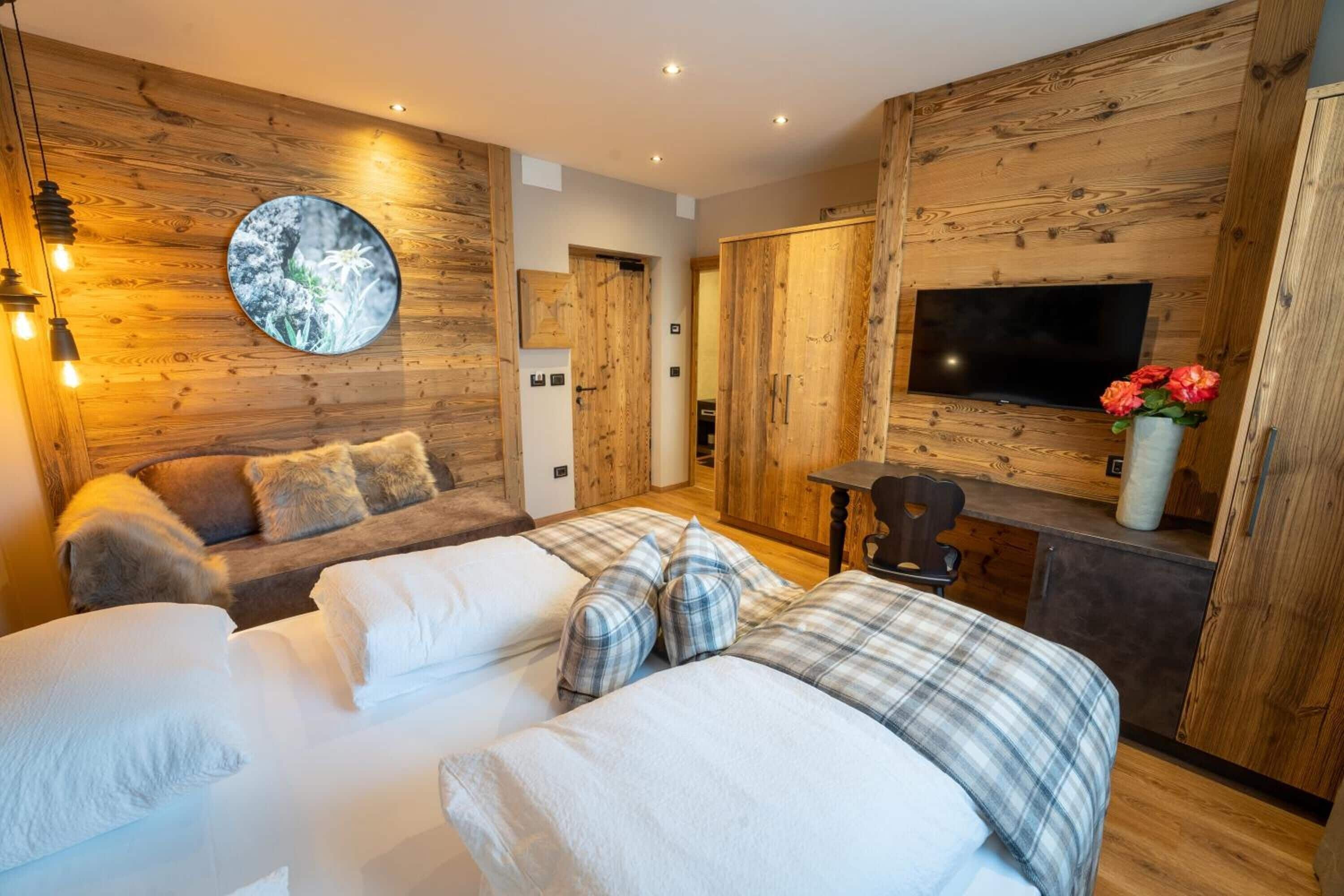 A cozy bedroom with a wooden wall, a round mirror, a bed with plaid bedding, a sofa with fluffy pillows, a TV, and a small table with a vase of flowers.