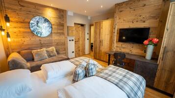 A cozy bedroom with a wooden wall, a round mirror, a bed with plaid bedding, a sofa with fluffy pillows, a TV, and a small table with a vase of flowers.