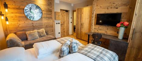 A cozy bedroom with a wooden wall, a round mirror, a bed with plaid bedding, a sofa with fluffy pillows, a TV, and a small table with a vase of flowers.